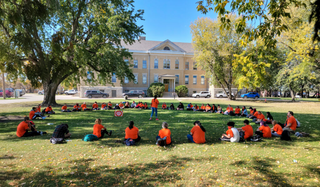 A large group of people, many wearing orange shirts, sit in a wide circle on a grassy campus lawn while a person stands speaking in the centre, with trees and a historic building in the background.