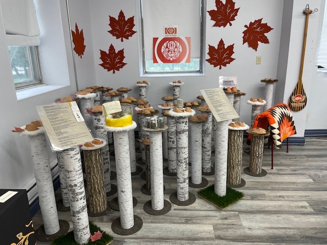 Indoor art installation of birch-log columns topped with handmade mushrooms, surrounded by red maple-leaf decorations and Indigenous artwork.