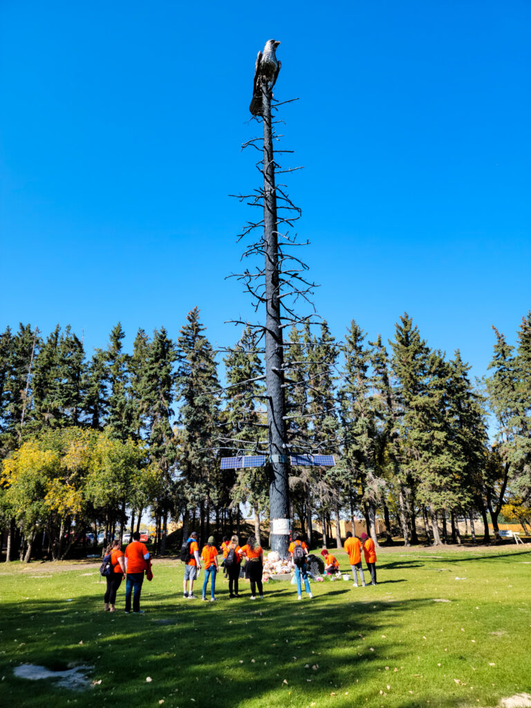 People wearing orange shirts gather around a tall memorial sculpture featuring an eagle perched atop a bare tree, with offerings placed at its base in a park.