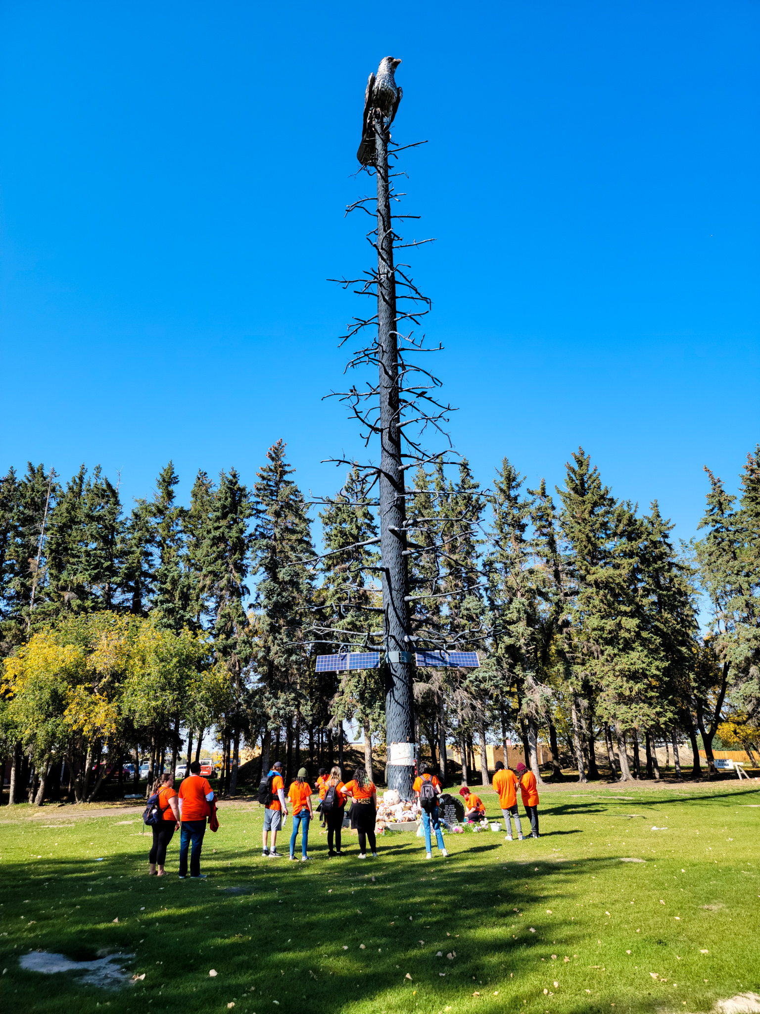 People wearing orange shirts gather around a tall memorial sculpture featuring an eagle perched atop a bare tree, with offerings placed at its base in a park.
